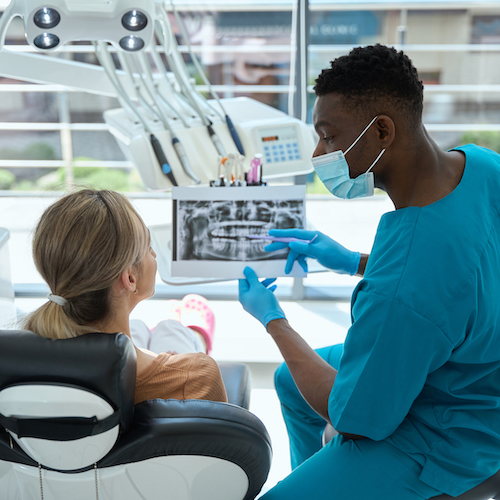 Young doctor in protective mask and blue suit sitting on chair near woman and showing results of treatment