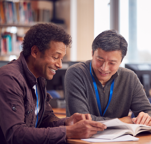 Teacher With Mature Male Adult Student Sitting At Table Working In College Library